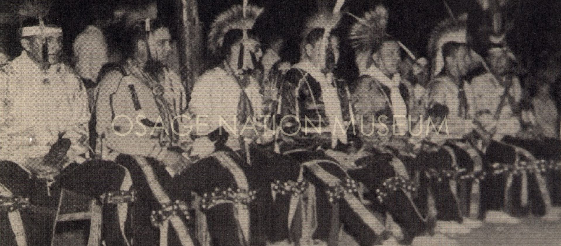 An important component of the June dances are the bells such as those worn by the following men seated under the dance arbor, L-R: Ed Beartrack, Francis Drexil, Jamison Bear Jr., Harold West, Clem Mason, Archie Mason, Ed Red Eagle.